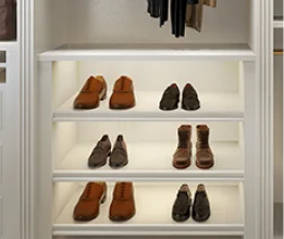 Backlit white wardrobe shelving displaying pairs of brown and black shoes with hanging clothes above